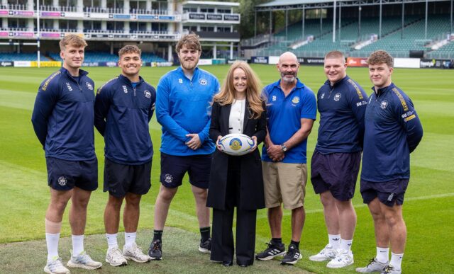 A group of rugby players, coaches and sponsors stand on the pitch at The Rec rugby stadium in Bath. The woman in the middle, Colour Studios Director Rowan Freeman, is holding a rugby ball with the University of Bath logo on it.