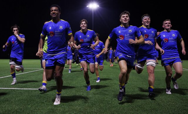 A group of University of Bath rugby players running towards the camera, under floodlights, with smiles on their faces.