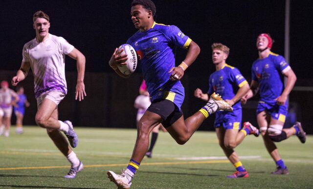 University of Bath rugby player Tyler Offiah, with ball in hand, smiles as he runs in for a try. Two of his team-mates, also wearing blue, are behind him.