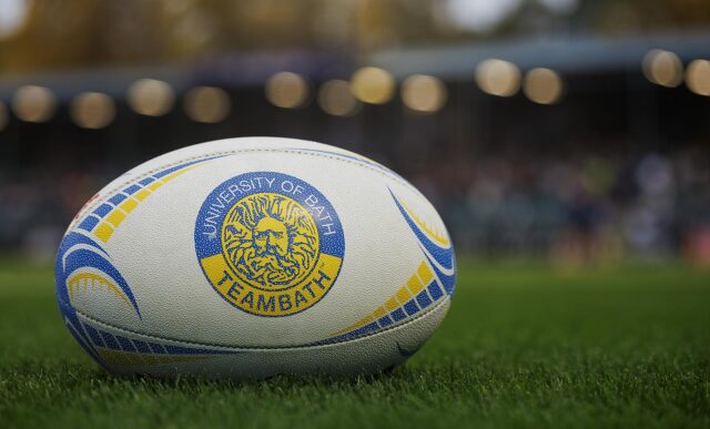 A close up of a rugby ball, branded with the Team Bath logo, on a pitch