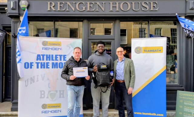 Enoch Opoku-Gyamfi, centre, holding a backpack. Either side of him are Rengen CEO Iestyn Lewis, holding an Athlete of the Month certificate, and University of Bath Director of Sport Stephen Baddeley. They are standing in front of the entrance to Rengen House and behind them are two pop-up banners, one saying "Athlete of the Month" and the other bearing the Team Bath logo.
