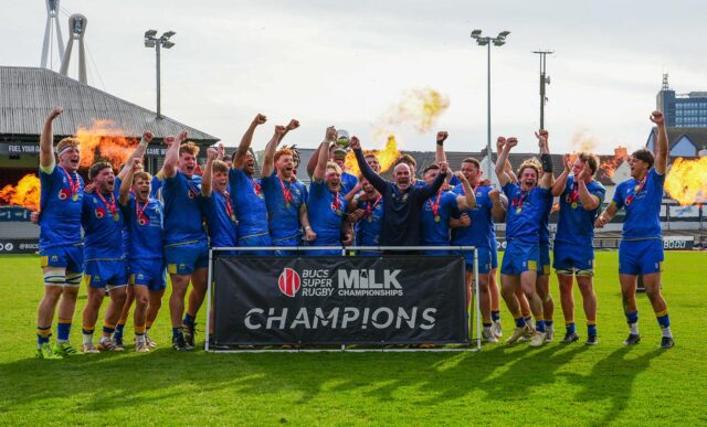Captain Max Pearce and Head of Rugby Aaron James lift the BUCS Super Rugby Milk Championship trophy as their team-mates celebrate and fireworks go off behind them
