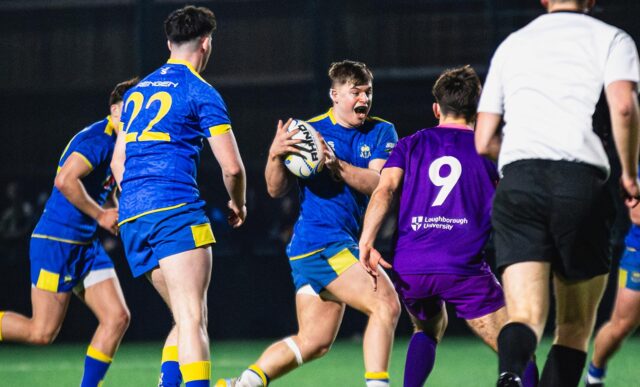A rugby player, wearing blue, running with the ball as a player in purple closes him down.