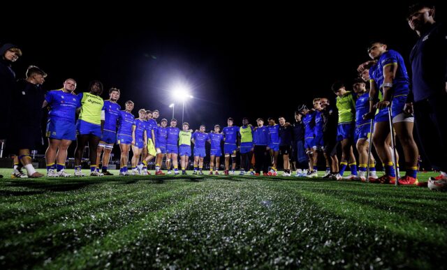 A group of rugby players and coaches in a huddle underneath floodlights